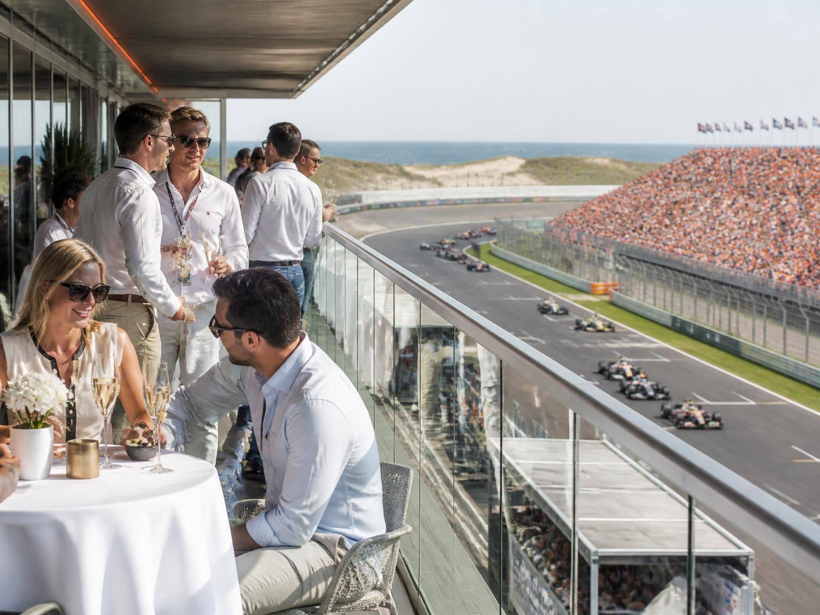 Gasten met champagne op het Paddock Club terras boven de pitboxen met zicht op de raceauto's
